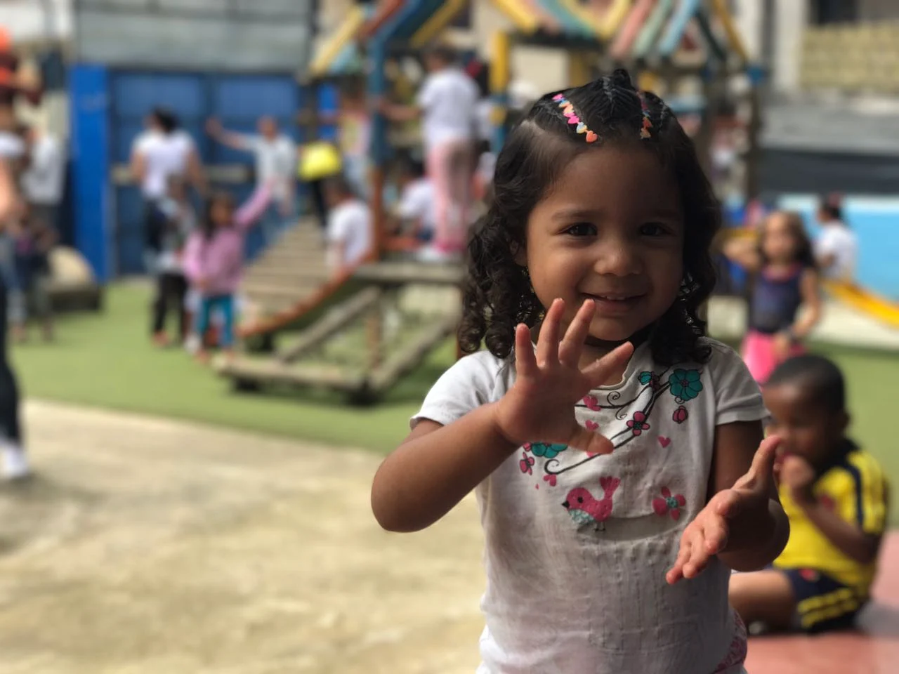 Niña en un espacio de recreación infantil, con juegos y otras personas al fondo.